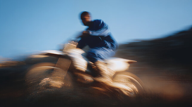 Male motorcyclist in blue outfit riding a dirt bike at high speed, creating a dynamic motion blur effect against a clear blue sky, showcasing adrenaline and excitement