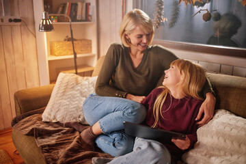 A mother sits close to her daughter on a comfortable couch, both sharing smiles and laughter while enjoying time together in a cozy living room setting.