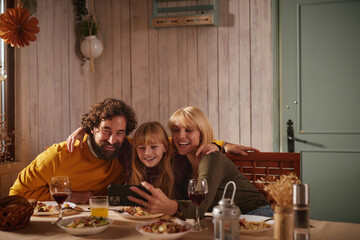 A family gathers around a table filled with autumn dishes. They share smiles and laughter while looking at a smartphone together in a warm, inviting space.