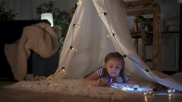 Child reading under tent with flashlight, girl lies on rug surrounded by string light, cozy blanket and book, indoor night play, smiling expression, warm glow illuminating face and imaginative focus.