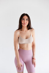 Fototapeta premium Sports woman standing in activewear with abs muscles, looking at camera isolated on white background. Caucasian 30s fitness girl wears beige top, pink leggings. Slim fit smiling brunette, blue eyes.