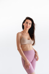 Fototapeta premium Sports woman standing in activewear with abs muscles, looking at camera isolated on white background. Caucasian 30s fitness girl wears beige top, pink leggings. Sexy fit smiling brunette, blue eyes.