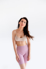 Fototapeta premium Sports woman standing in activewear with abs muscles, looking at camera isolated on white background. Caucasian 30s fitness girl wears beige top and pink leggings. Slim fit brunette with blue eyes.