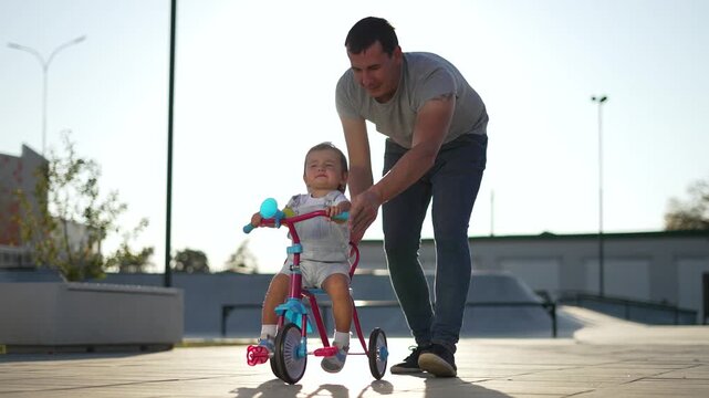 Man teaches toddler to ride tricycle while father and dad encourage child learning balance parent support outdoor near skatepark with interaction and steady hand guiding confident pedal practice
