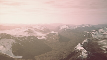 Mountains stretch under a delicate layer of snow as the sunset casts a pink glow over the landscape. Valleys below reveal rich hues and vastness, inviting exploration and wonder.