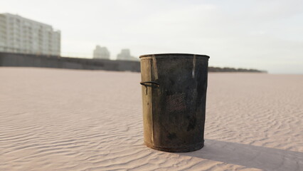 Naklejka premium A weathered trash can stands alone on a serene beach, surrounded by soft sand. In the background, city buildings rise against a beautiful sunset sky, creating a peaceful atmosphere.