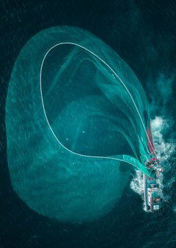 Aerial view of a fishing vessel dragging a large net through the turquoise waters, creating a swirling pattern visible from above, Tuy Hoa, Phu Yen, Vietnam.