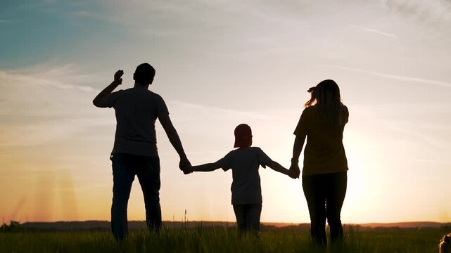 Holding hands together at sunset family silhouette of parent child and dog walking across grassy field with warm sky and horizon, parent and child holding hand in golden glow and long shadow