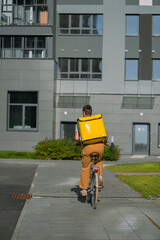 Naklejka premium Bike courier riding bicycle carrying yellow insulated delivery backpack, delivering food or groceries in city, working in urban environment, freelance worker on gig economy