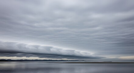 A wide gray sky with a low shelflike cloud formation hangs over a still reflective beach and distant mountains