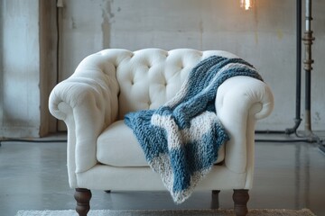 A comfortable cream tufted armchair features a chunky blue and off-white knit blanket, creating a cozy corner in an industrial setting.