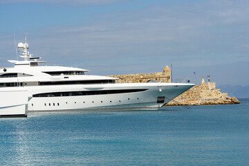 Luxury white superyacht moored by historic stone fortress in calm turquoise sea, symbol of wealth, tourism and nautical lifestyle under soft cloudy sky.