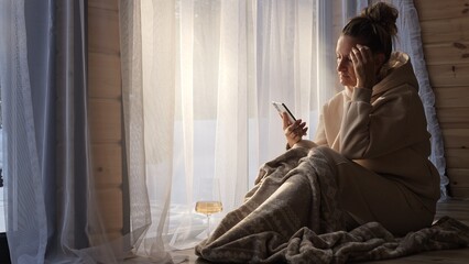 Woman wrapped in blanket near window holds wine and scrolls phone showing digital reflection...