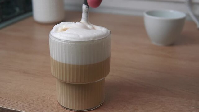 close-up of a spoon stirring the thick foam of a layered latte or cappuccino in a modern ribbed glass cup on a wooden table 