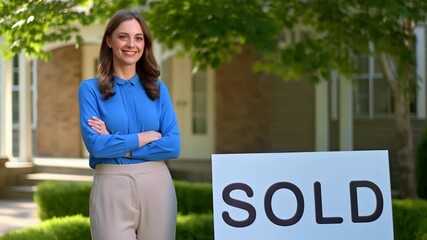 Smiling female real estate agent standing outdoors, arms folded, beside a sold sign in front of a residential house, representing successful property sales