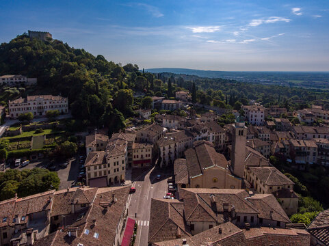 Aerial view of terracotta rooftops cascading toward the Duomo, shadowed by the Rocca fortress atop the verdant hill, under a serene sky, Asolo, Veneto, Italy.