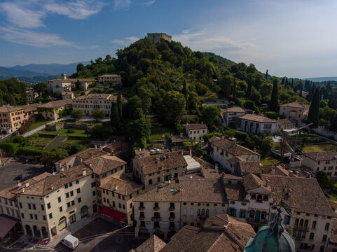 Aerial view of terracotta rooftops cascade down towards ancient buildings, crowned by the distant Rocca fortress atop a verdant hill, Asolo, Veneto, Italy.