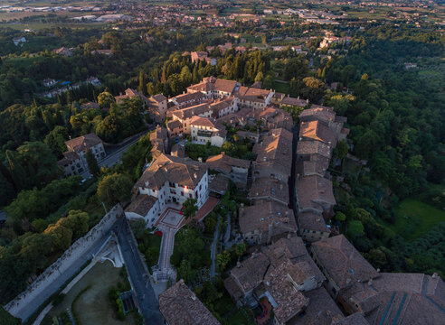 Aerial view of terracotta rooftops blending into the verdant hillsides, the ancient walls of the town embrace the sunlit buildings in a warm, golden glow, Asolo, Veneto, Italy.