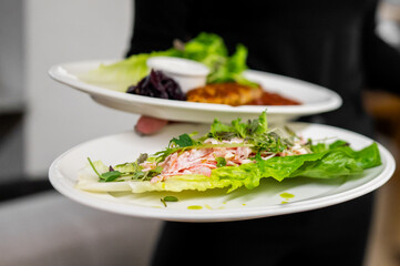 A server presents two plates of contemporary, healthy cuisine. The front plate showcases delicate lettuce wraps filled with a pink salad mix and microgreens, drizzled with oil.