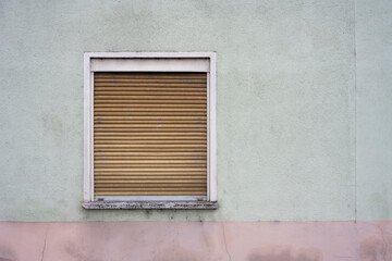 Dirty old wall, painted in green and pink, brown wooden jalousie rolled up, window with closed shutter