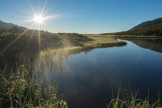 View of sunlight bursts across a tranquil lake reflecting the clear sky, fringed by green vegetation and distant mountains, Biele pleso tarn, Presovsky kraj, Slovakia. - Powered by Adobe
