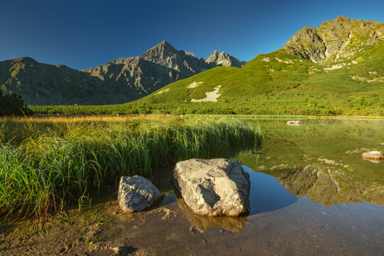 View of jagged peaks pierce the azure sky above a tranquil lake reflecting the surrounding vibrant green landscape, Biele pleso tarn, Vysoke Tatry, Presovsky kraj, Slovakia.