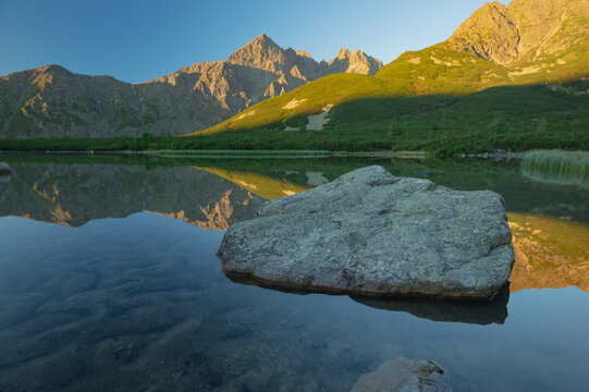 View of mirrored peaks meet tranquil waters, granite stone rises from the depths under the golden kiss of dawn, Biele pleso tarn, Vysoke Tatry, Presovsky kraj, Slovakia.
