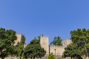 Naklejka premium The impressive medieval stone walls and battlements of the Castle of Torres Novas, Portugal, rise above a foreground of lush green trees.