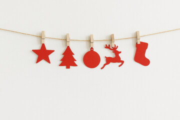 Red wooden Christmas ornaments hanging on a string with clothespins on a clean white background, minimal festive decor featuring star, tree, bauble, reindeer and stocking shapes