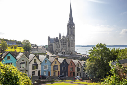 View of a majestic cathedral towers over a vibrant row of colorful houses, set against a backdrop of shimmering water, Cobh, County Cork, Ireland.