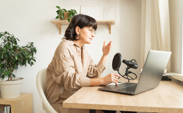 Mature caucasian woman podcasting from home office with laptop and microphone setup - Powered by Adobe