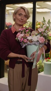 Vertical shot of middle-aged Caucasian woman holding hatbox bouquet of flowers and looking at camera with smile in florist shop