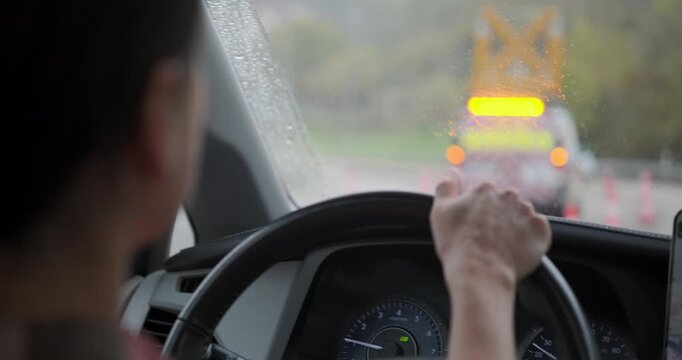 Over the shoulder view of a woman driving a car in rainy weather and slow traffic. Hands on steering wheel, wipers clearing the glass, construction lights visible ahead. Slow motion.