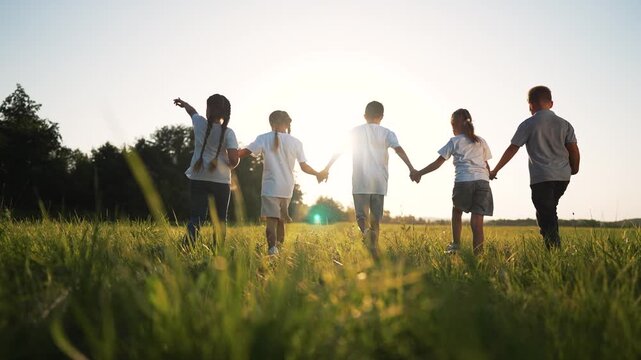 Children holding hand walk across sunlit field with tall grass at sunset while friend and kid play with soccer ball showing friendship joy and carefree child under golden light near tree line