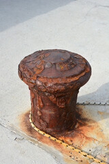Fototapeta premium A heavily rusted, cylindrical metal mooring bollard stands on a concrete pier, with a thick, frayed rope looped around its base