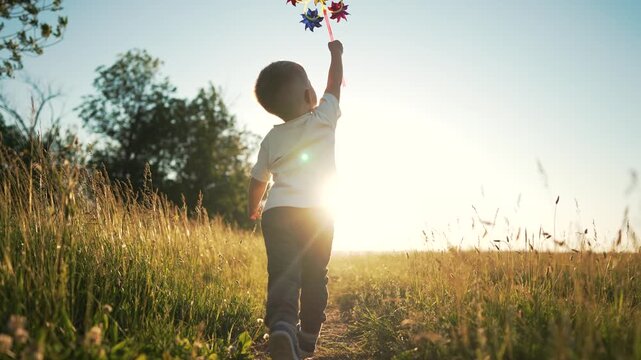 Running boy holds colorful pinwheel across sunlit grass field at sunset, child walking toward horizon with backlit sun silhouette, summer outdoor play in meadow, carefree energy and golden light - Powered by Adobe
