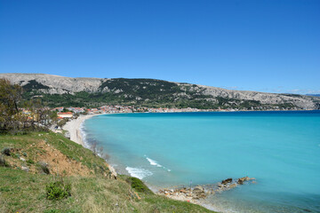 Obraz premium A panoramic view captures the crescent-shaped Vela Plaza beach and the town of Baska on Krk Island, with turquoise sea and a dramatic mountain backdrop under a clear sky