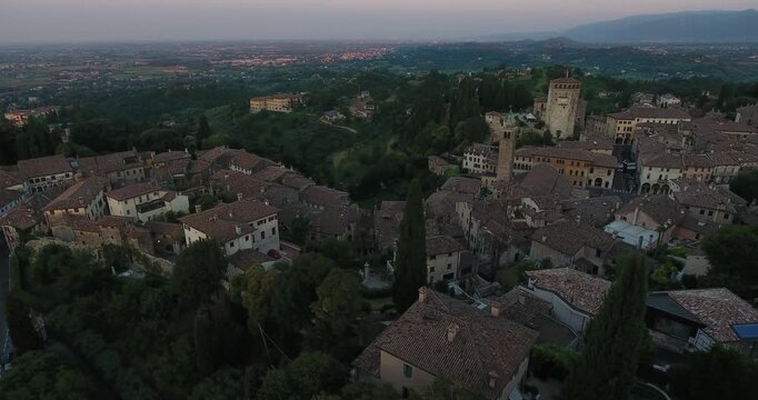 Aerial view of Asolo's dark, tiled rooftops nestling among lush green trees under a dusky sky, creating a tapestry of textures, Asolo, Veneto, Italy.