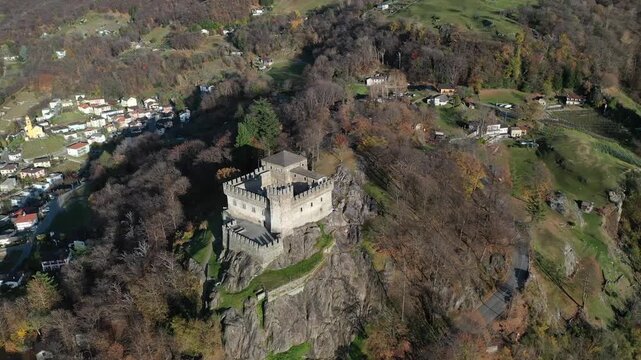 Aerial view of the historic Sasso Corbara castle perched atop a rocky hill, surrounded by autumn foliage in golden and brown hues, Bellinzona, Ticino, Switzerland.