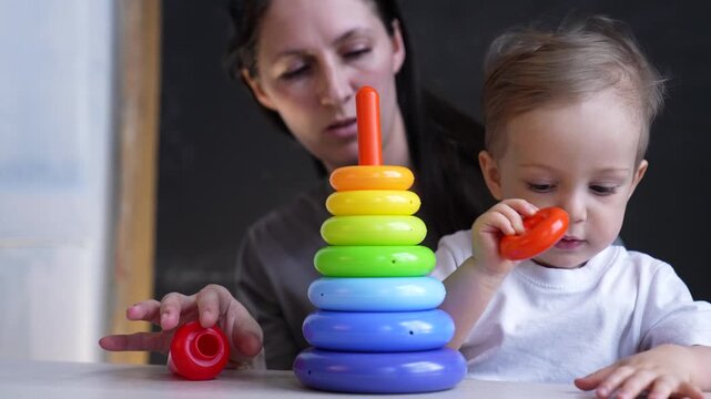 Baby stacks colorful ring toy while mother guides toddler through play and learning activity, child focused on rainbow stacking ring and parent supporting motor development indoors with toy