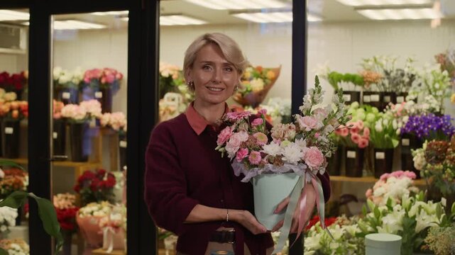 Medium portrait of positive Caucasian middle-aged woman holding hatbox bouquet and smiling at camera in florist shop