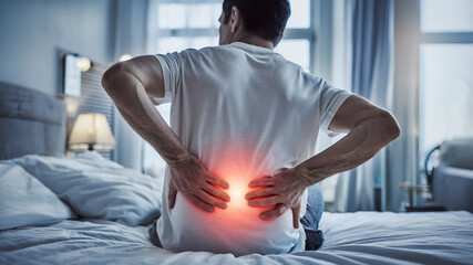 A man suffering from lower back pain, touching muscle inflammation on his bed, lumbar red warning highlight