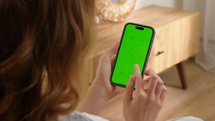 Woman sit use digital tech cell phone with green screen mock up. Hand scroll phone web site, swipe like photos. Girl hold mobile closeup. Chroma key page. Cozy home background. Online app copy space.