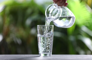 Woman pouring water into glass from jug at grey table against blurred green background, closeup. Space for text