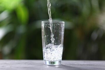 Pouring water into glass at grey table against blurred green background, closeup