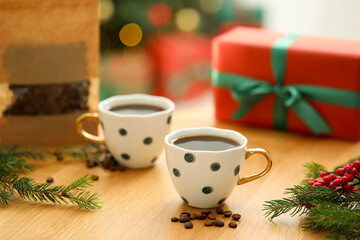 Aromatic coffee in cups, fir tree branches, beans and gift box on wooden table against blurred lights, closeup. Christmas greeting card