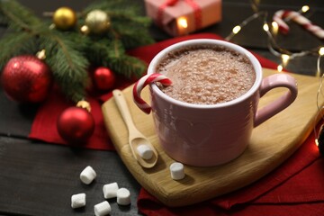 Tasty hot cocoa drink with candy cane in cup, marshmallows and Christmas decor on dark wooden table, closeup