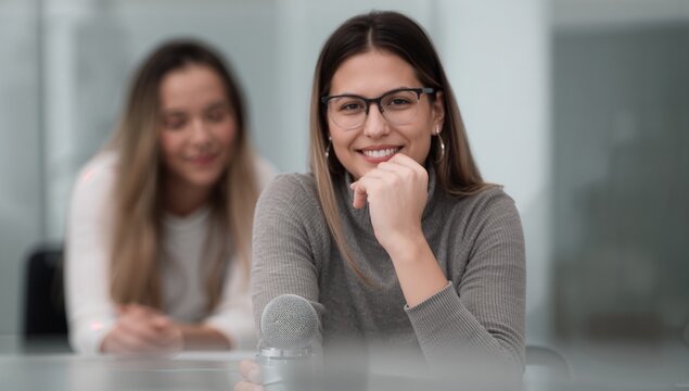 Confident young woman with glasses smiles at camera, ready to record podcast or interview in modern studio with colleague blurred behind.