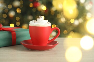 Aromatic coffee with whipped cream and Christmas gift on white wooden table against blurred lights, closeup