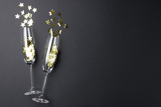 Merry Christmas and Happy New Year. Flute glasses with golden star shaped confetti on black background, flat lay. Space for text - Powered by Adobe
